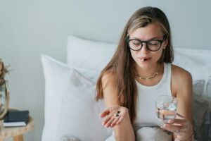 Concerned young woman with glasses holds pills and water, considering health in comfortable bedroom