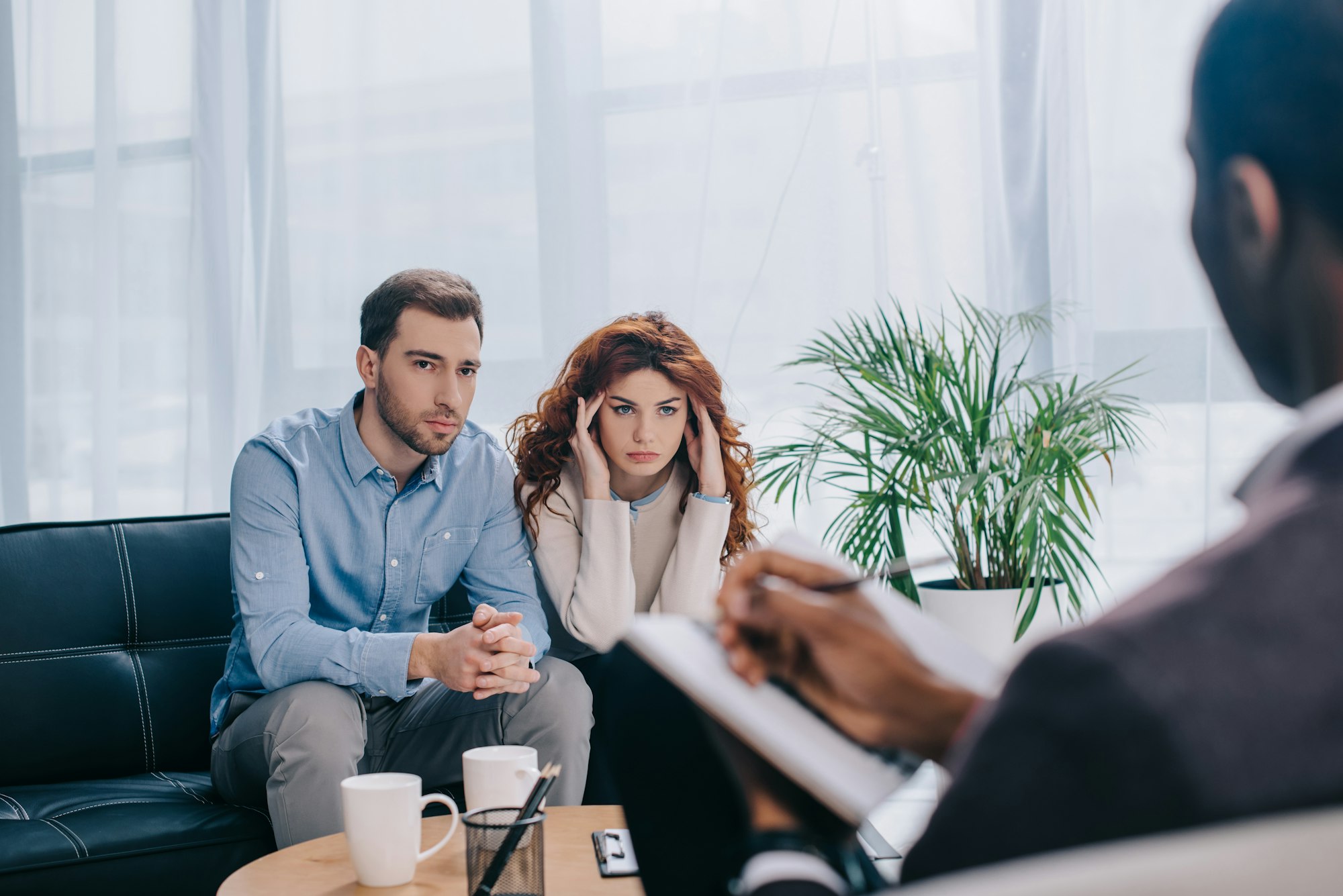 Upset young woman and boyfriend on sofa and counselor with textbook