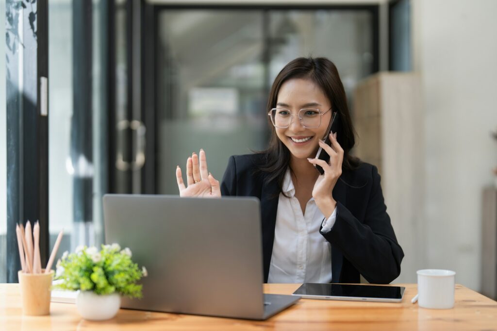 Asian businesswoman talking with client on mobile phone and working on laptop.