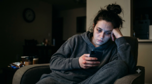 An exhausted woman slouching in a dark armchair late at night, staring blankly at her illuminated smartphone screen