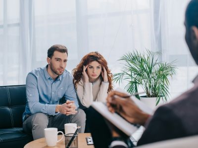 Upset young woman and boyfriend on sofa and counselor with textbook