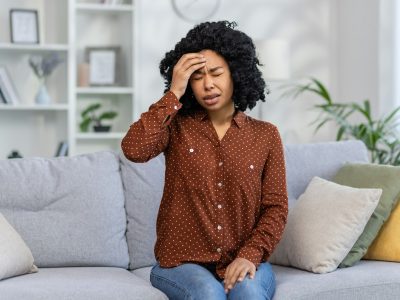 Worried young woman sitting on couch at home feeling stress, anxiety or headache